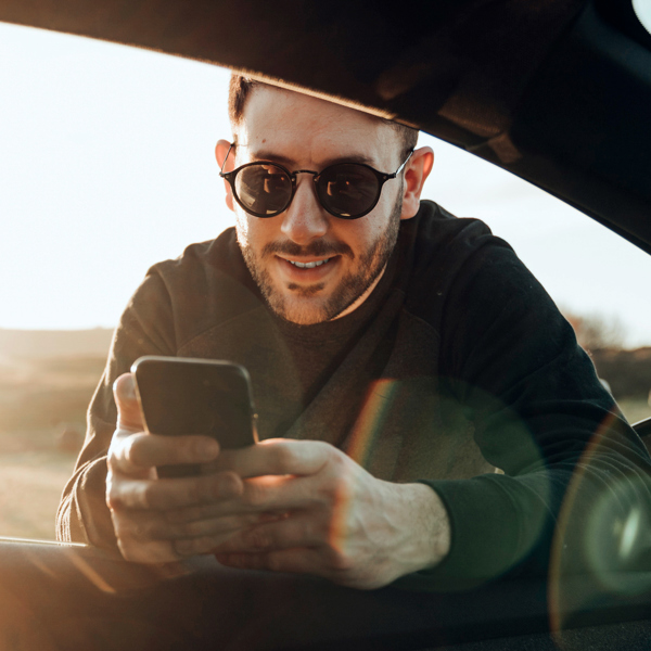 Kille med solglasögon lutar sig in i bil med mobil i handen i solnedång. Foto. 