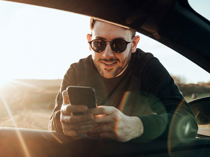 Ung man med solglasögon hänger in genom rutan till förarsätet i en bil, med sin mobiltelefon i händerna