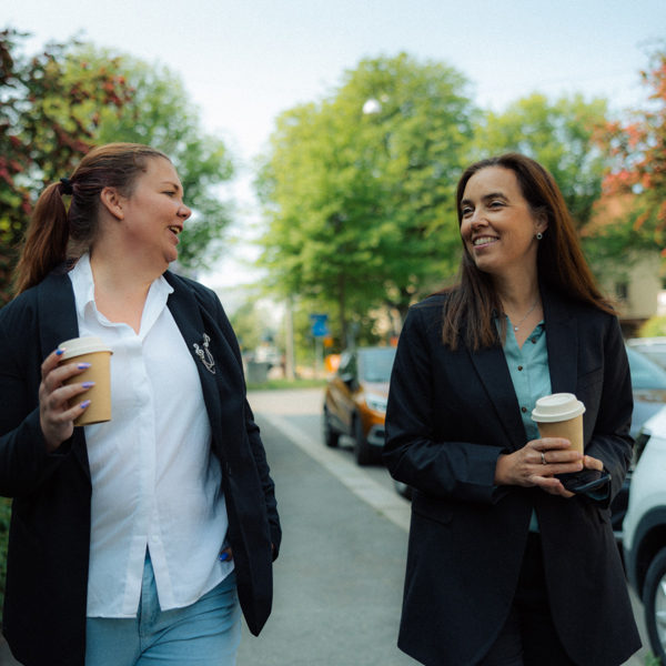 Två kvinnor ler mot varandra, de bär på varsin takeaway-mugg.